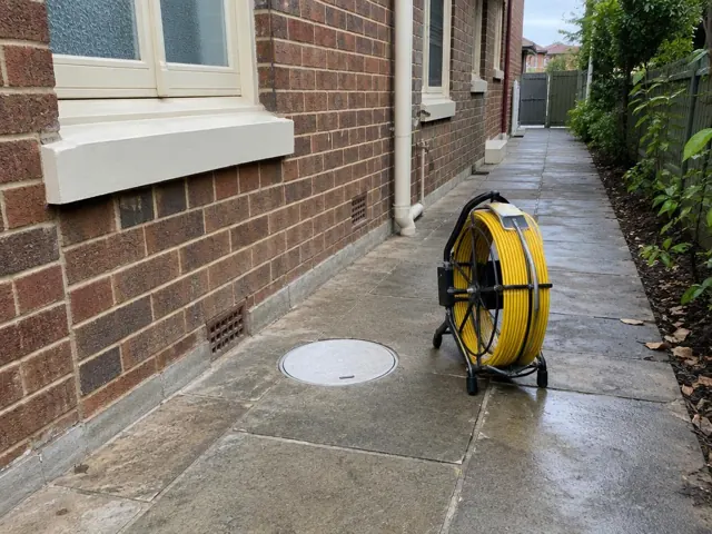 Capped drain inspection opening beside a yellow camera reel on a tiled side path at a brick house in Ryde