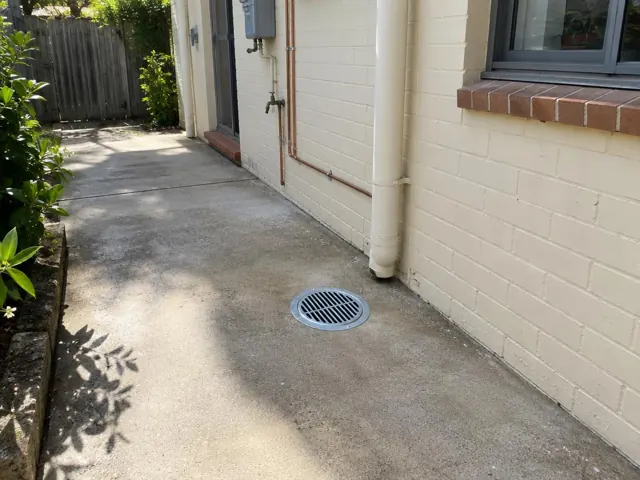 Outdoor floor gully with metal grate on a concrete side path beside a cream brick wall in Putney